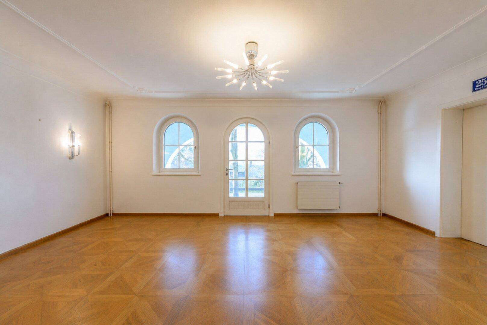Lichtdurchfluteter Raum mit Parkettboden, Stuckdecke und mehreren hohen Bogenfenstern.