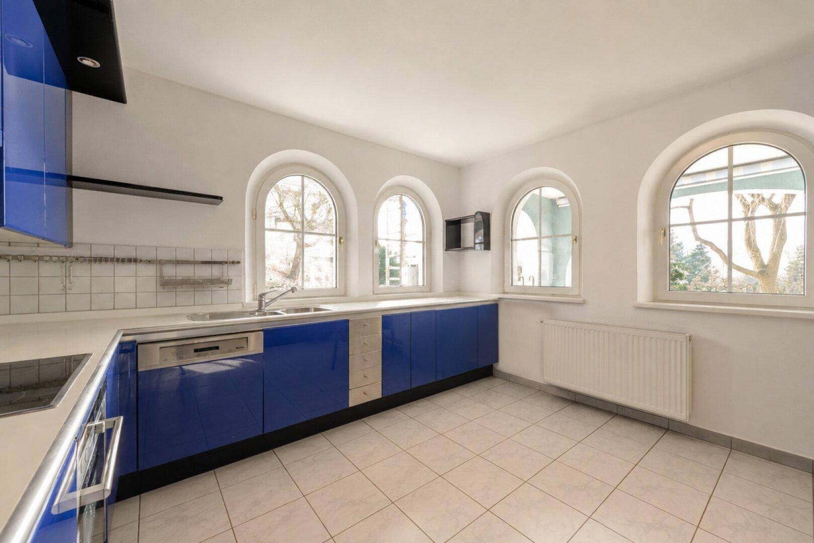 Moderne Küche mit blauen Hochglanzfronten, Fliesenboden und mehreren Bogenfenstern.