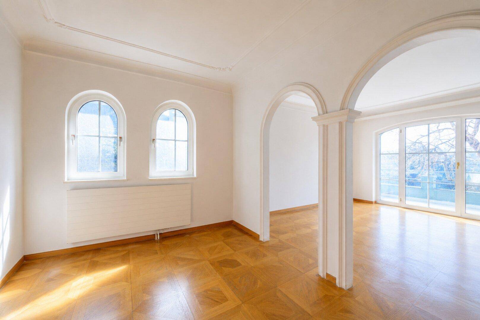 Eleganter Raum mit Parkettboden, Stuckdecke, Bogenfenstern und dekorativen Säulen.
