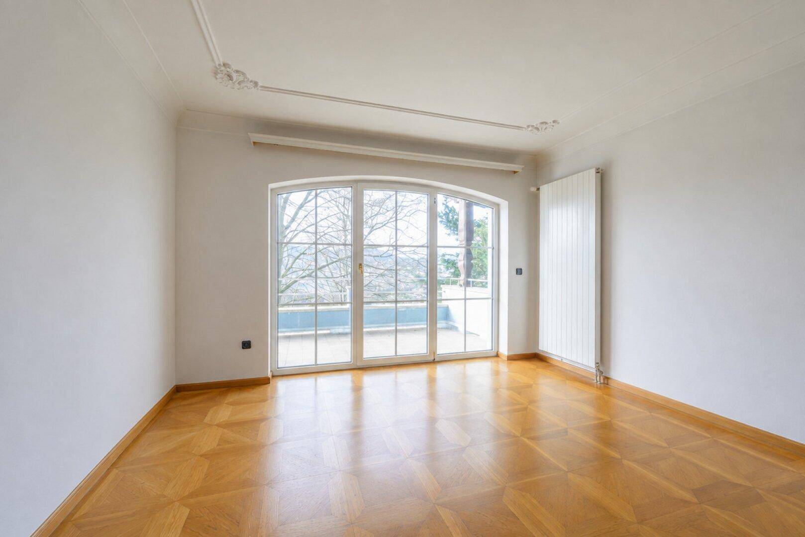 Heller Raum mit Parkettboden, Stuckdecke und großer Bogenfensterfront mit Zugang zur Terrasse.