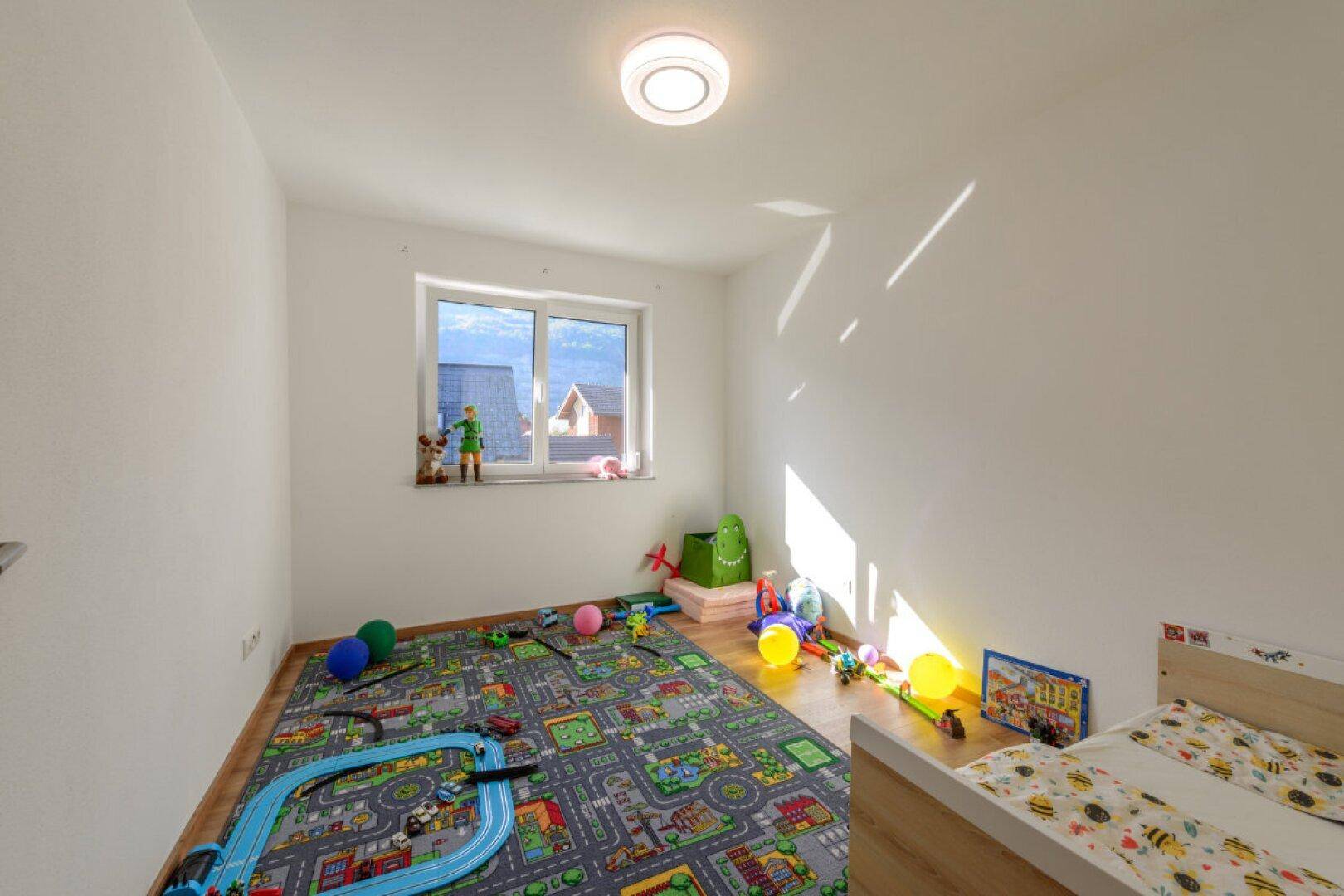 Helles Kinderzimmer mit Spielteppich, Bett und viel Tageslicht.