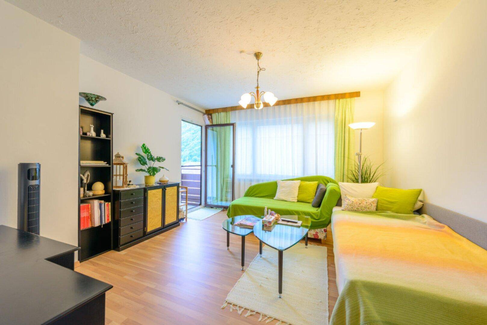 Helles Wohnzimmer mit grünem Sofa, Parkettboden und Zugang zu einem Balkon mit Bergblick.