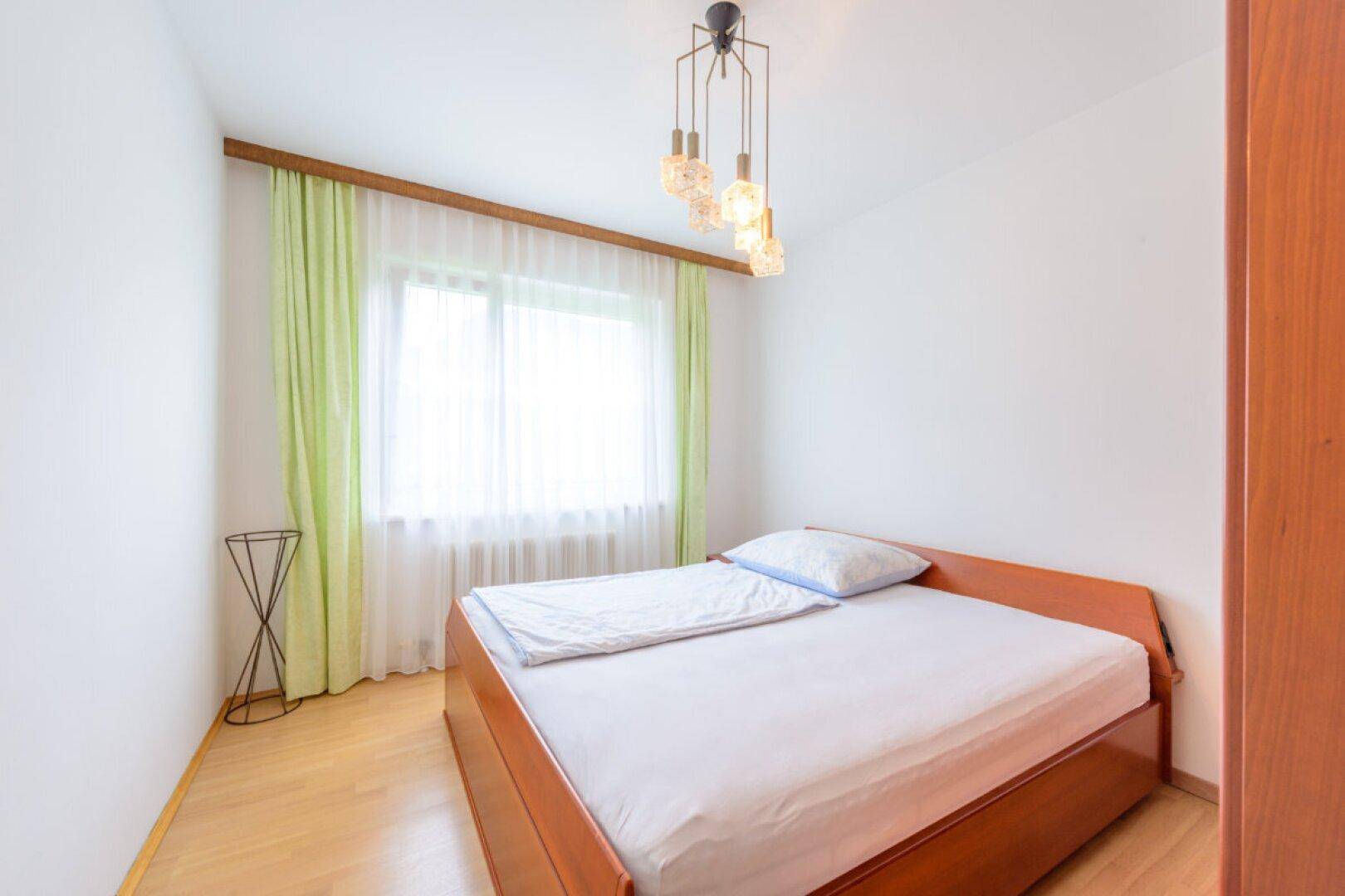 Helles Schlafzimmer mit Holzbettrahmen, weißen Wänden und Fenster mit hellgrünen Vorhängen.