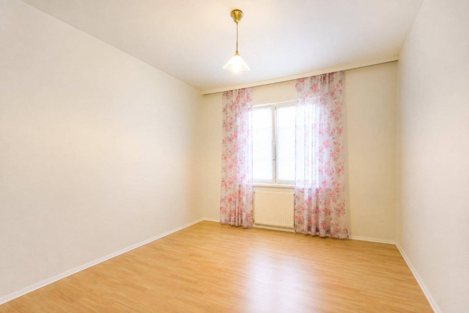 Leeres Zimmer mit Laminatboden und einem Fenster, das viel natürliches Licht hereinlässt.