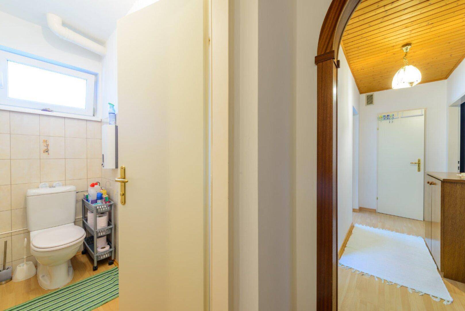 Separates WC mit Toilette, kleinem Fenster und hellen Fliesen, vom Flur aus zugänglich.