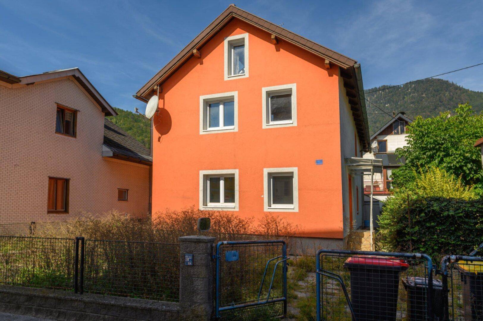 Vorderansicht des Wohnhauses mit oranger Fassade, Einfahrtstor und Mülltonnen.