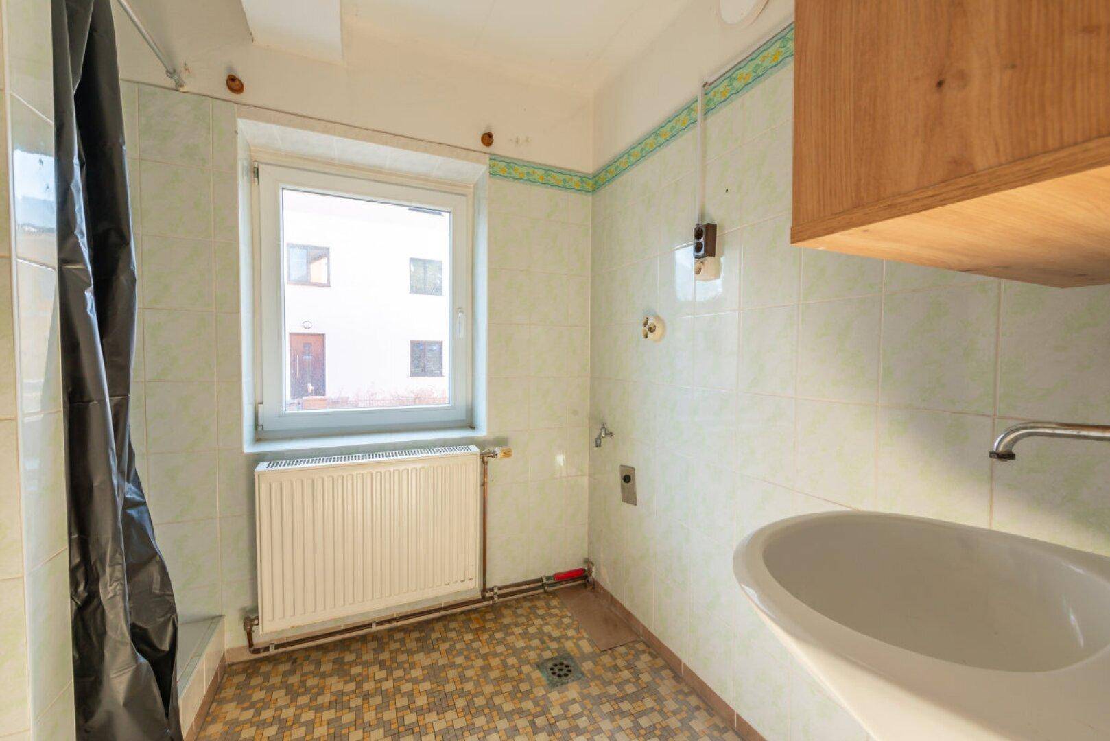 Badezimmer mit hellgrünen Fliesen, Waschbecken, Heizkörper und Fenster, das eine Modernisierung benötigt.