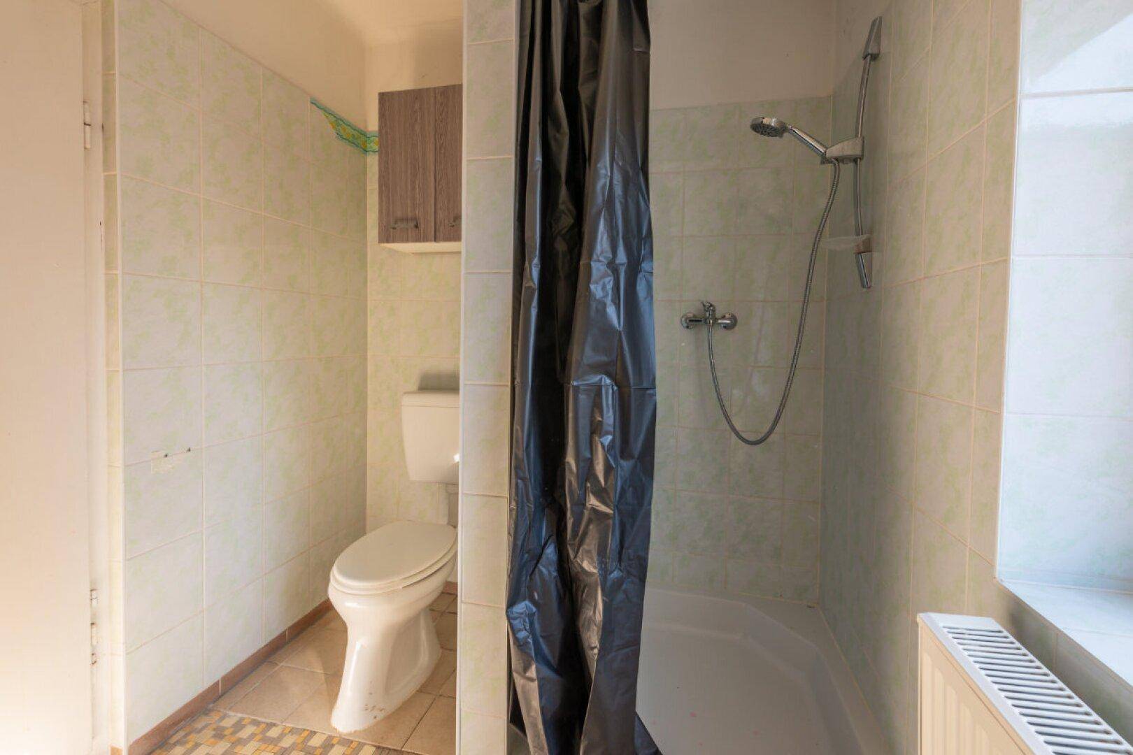 Badezimmer mit Duschbereich, WC und hellgrünen Fliesen, das eine Renovierung erfordert.