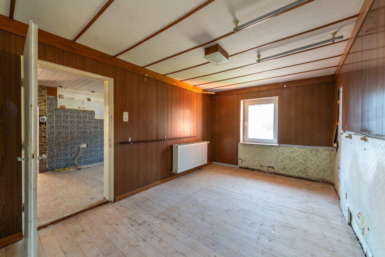 Zimmer mit Holzvertäfelung, Holzboden und Fenster, das auf eine Modernisierung wartet.