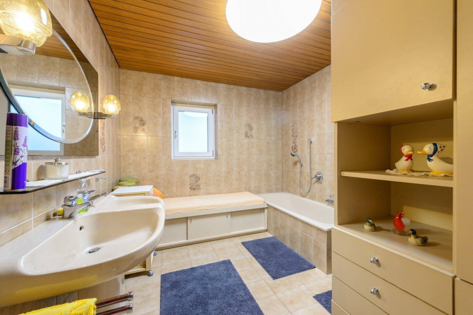 Badezimmer mit beigefarbenen Fliesen, Badewanne, Waschtisch und Holzdecke.