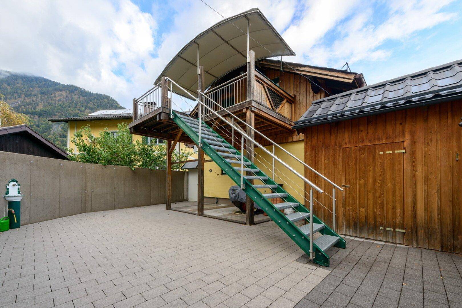 Außentreppe führt zu einer oberen Terrasse, umgeben von gepflastertem Hof und Holzschuppen.