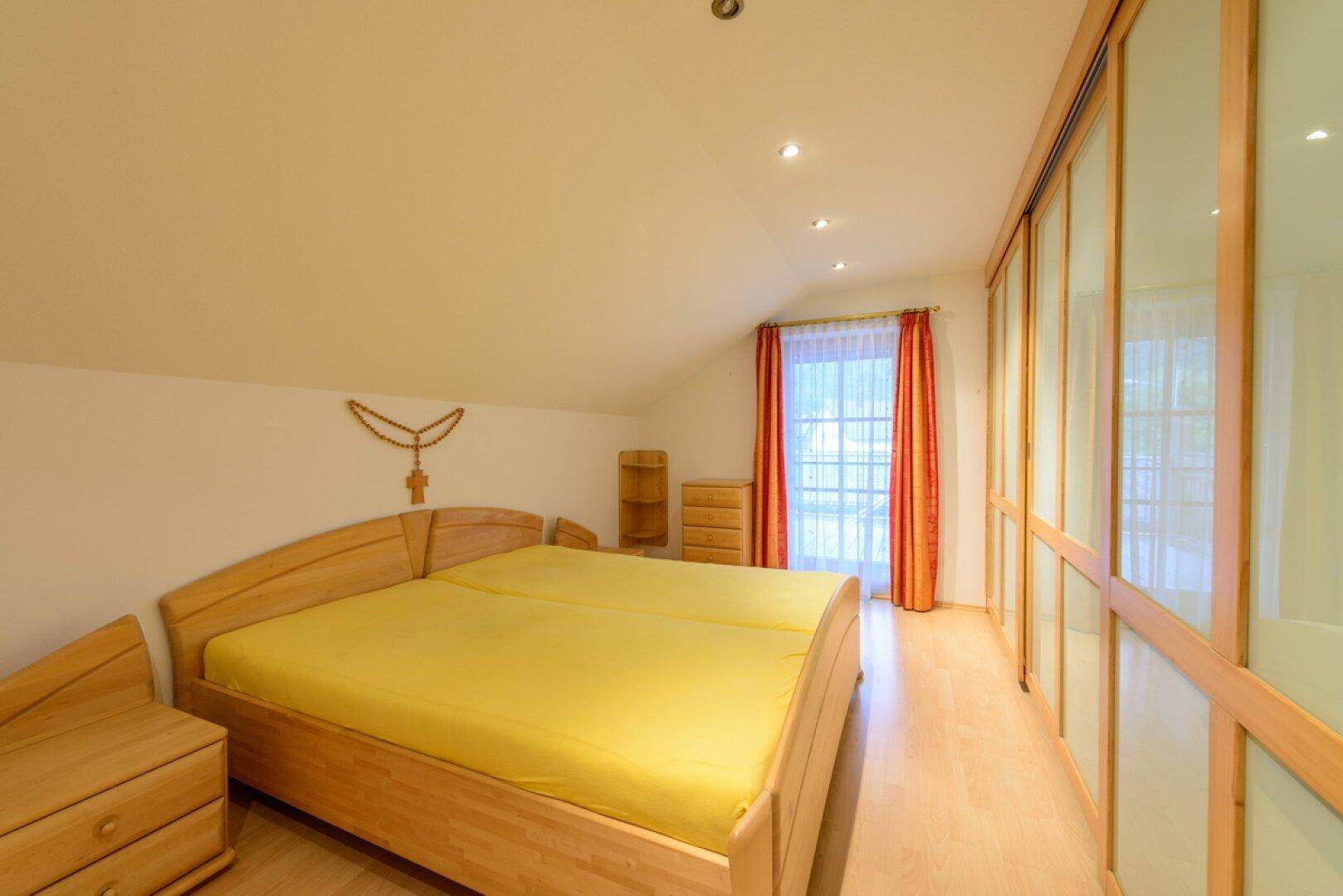 Schlafzimmer mit Holzbett, Einbauschränken und Dachschräge, hell durch Fenster belichtet.