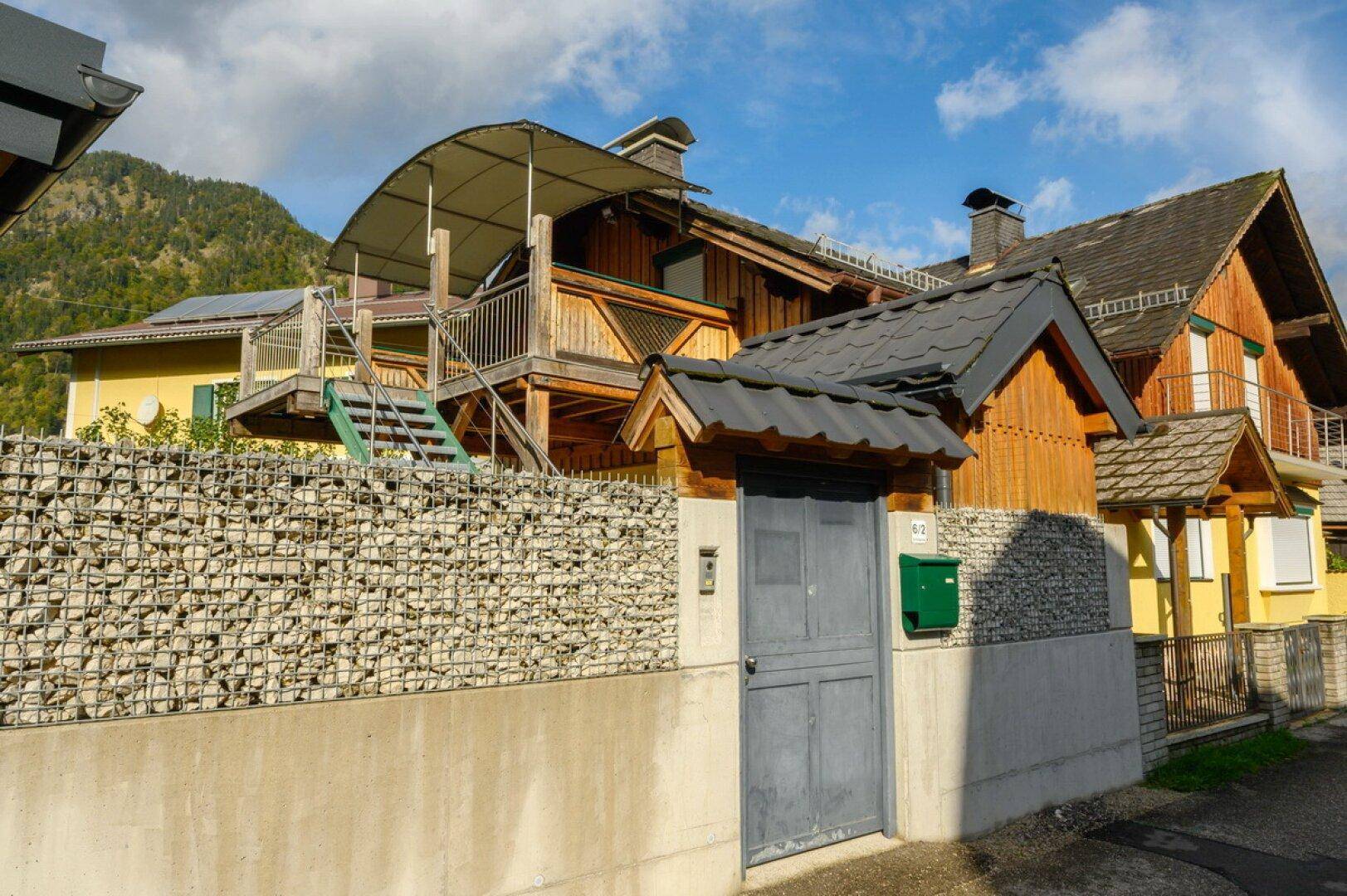 Ansicht des Hauses mit Gabionenmauer, Eingangstor und Außentreppe zum oberen Balkon.