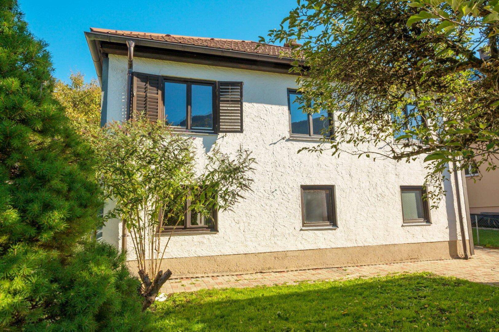 Seitenansicht eines Einfamilienhauses mit weißer Fassade, braunen Fensterläden und Garten.