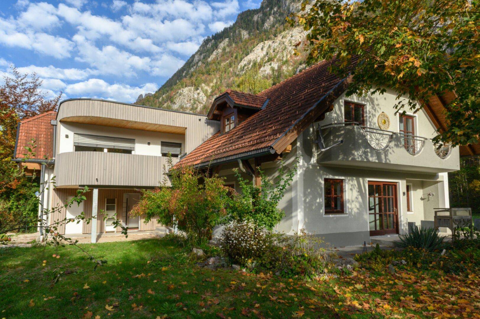 Hausfassade mit Balkonen und Holzelementen, eingebettet in eine grüne Landschaft mit Bergen.