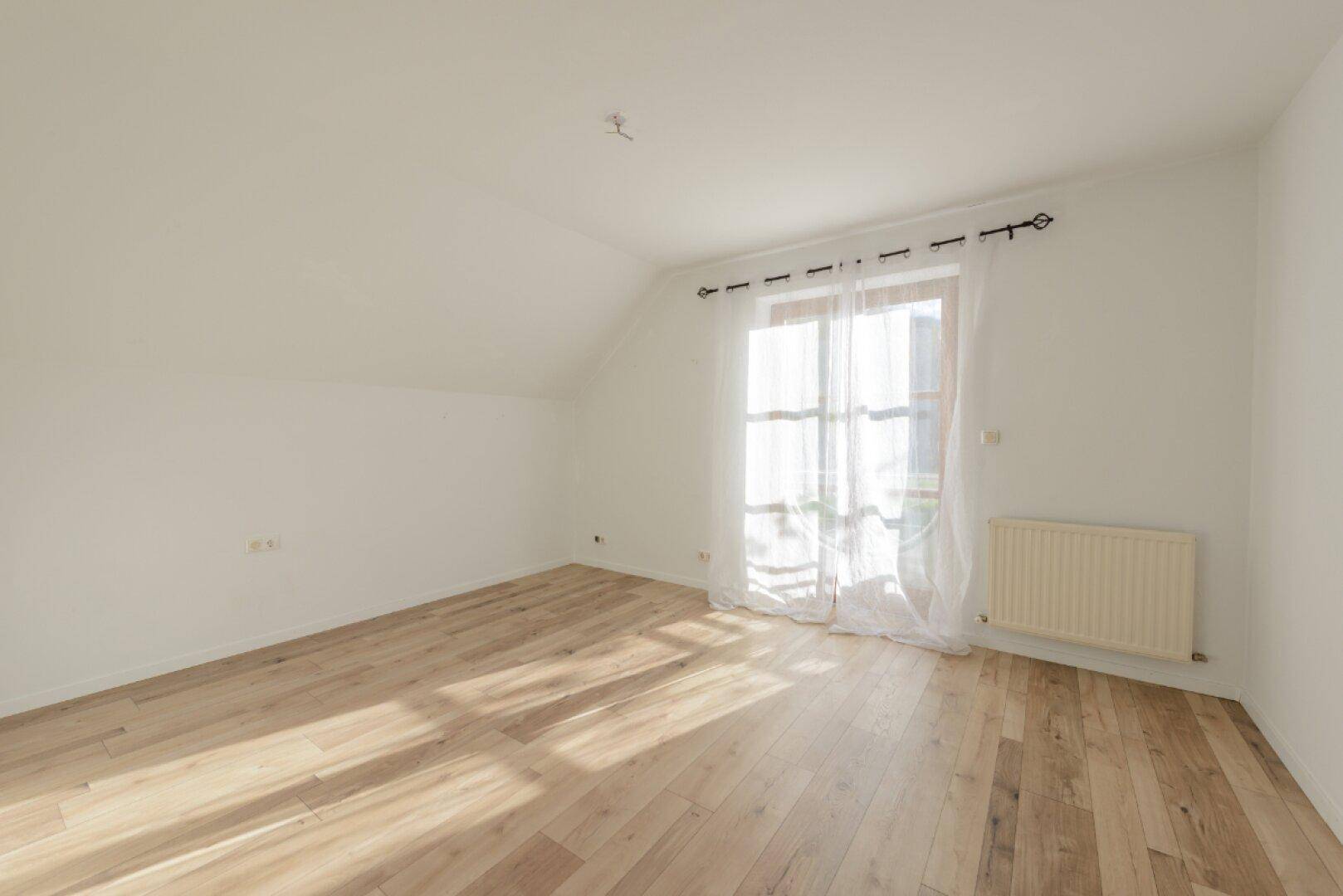 Helles Zimmer mit Holzboden, Dachschräge und großem Fenster mit viel Tageslicht.
