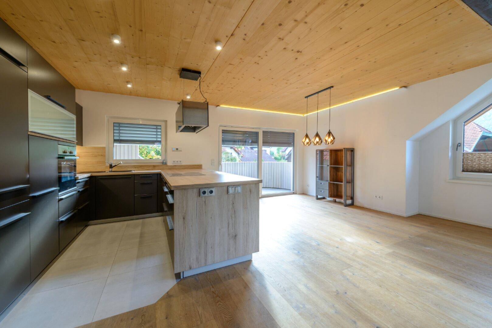 Moderne Küche mit dunklen Fronten, Holzdecke und Kücheninsel, offen zum Wohnbereich.