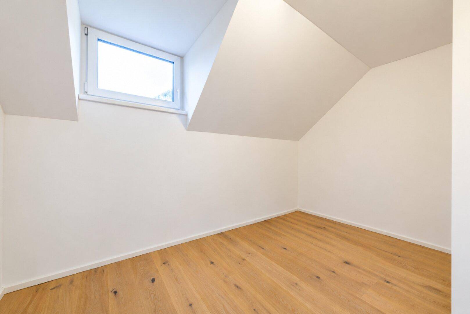 Kleines, helles Zimmer mit Holzboden und Dachfenster unter einer Dachschräge.