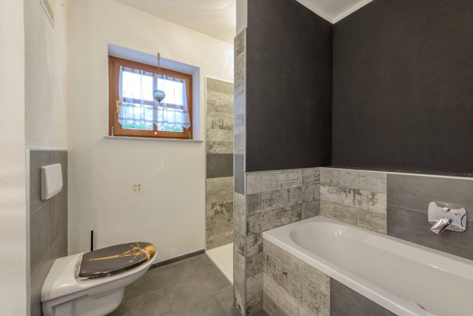 Modernes Badezimmer mit Badewanne, Dusche, WC und Fenster in Grautönen.