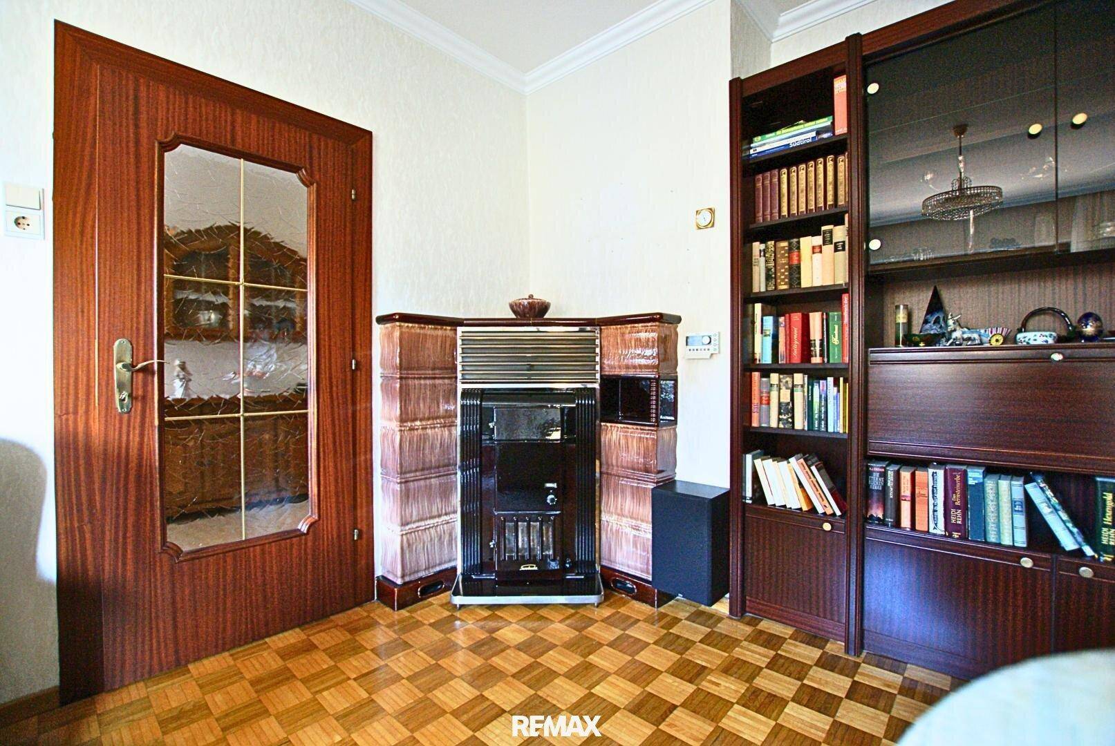 Wohnzimmer mit Kachelofen, Bücherregal und Holzboden, strahlt Gemütlichkeit aus.