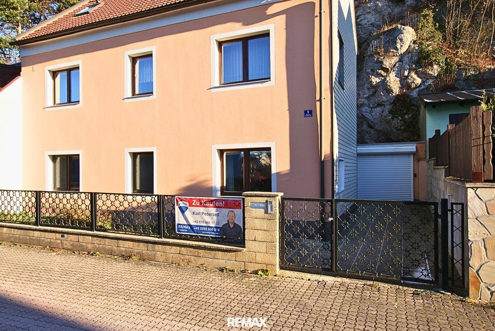 Ansicht des Hauses von der Straße mit dekorativem Zaun und Werbeschild, umgeben von Natur.