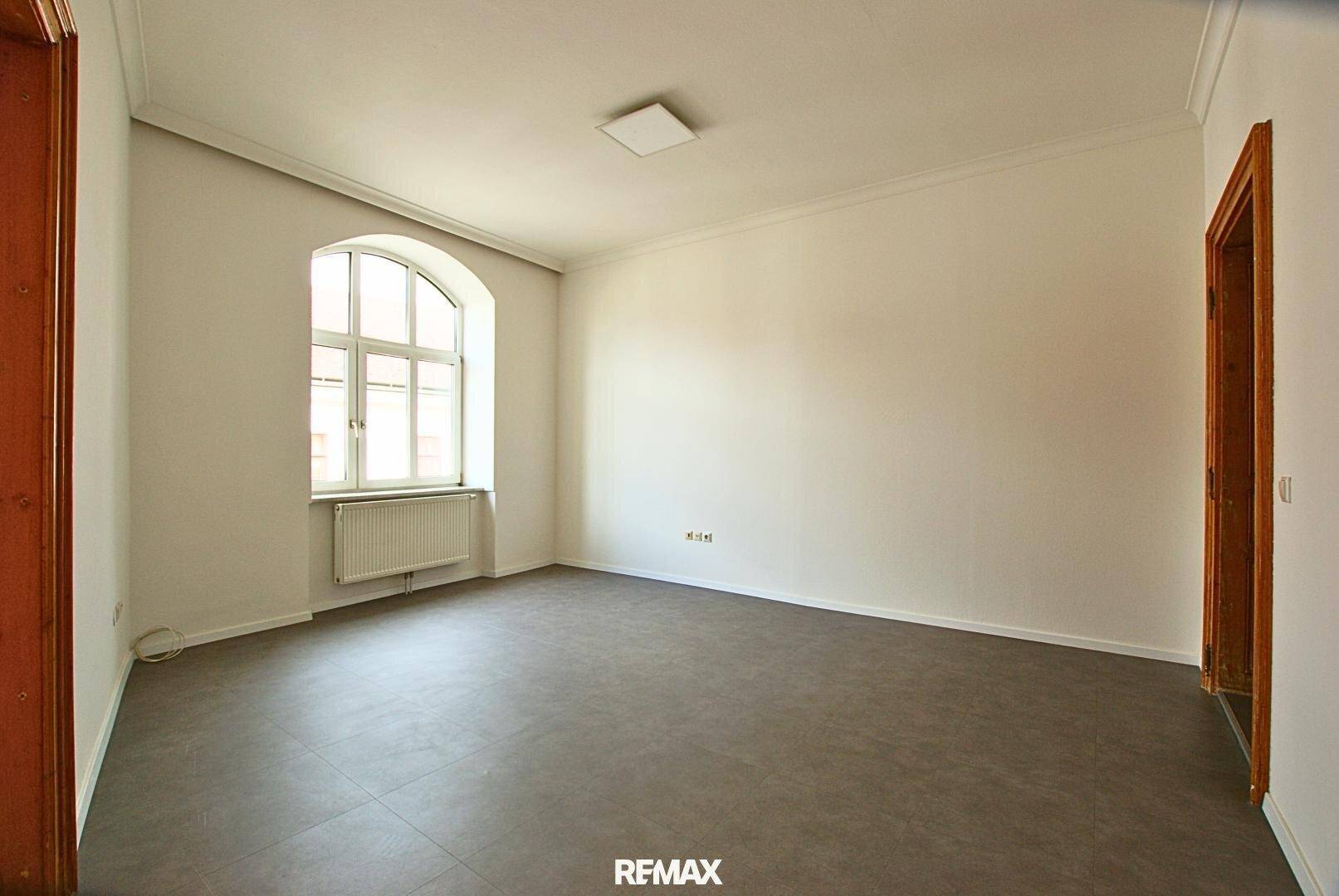 Leerer Raum mit hellen Wänden, grauen Fliesen und einem großen Bogenfenster mit Heizkörper.