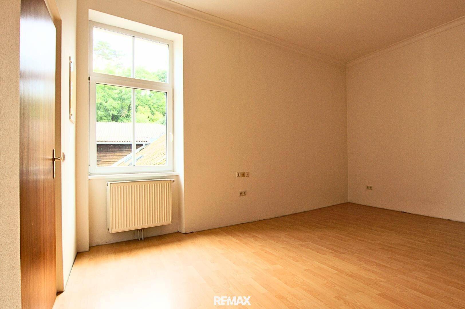 Leerer Raum mit Holzboden und einem Fenster, das einen Blick ins Grüne bietet.