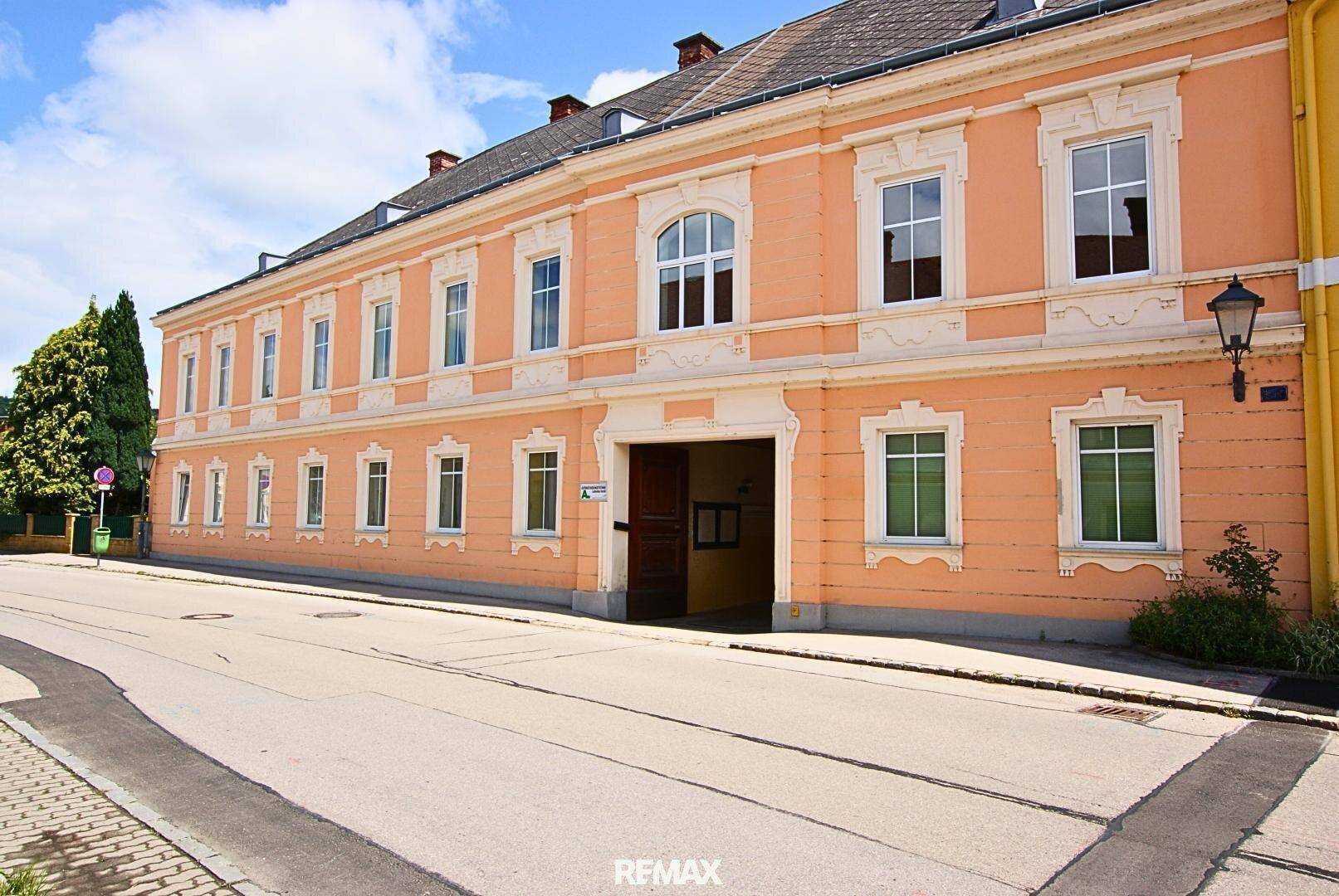 Historisches Gebäude mit oranger Fassade, vielen großen Fenstern und einem eleganten Torbogen an der Straße.