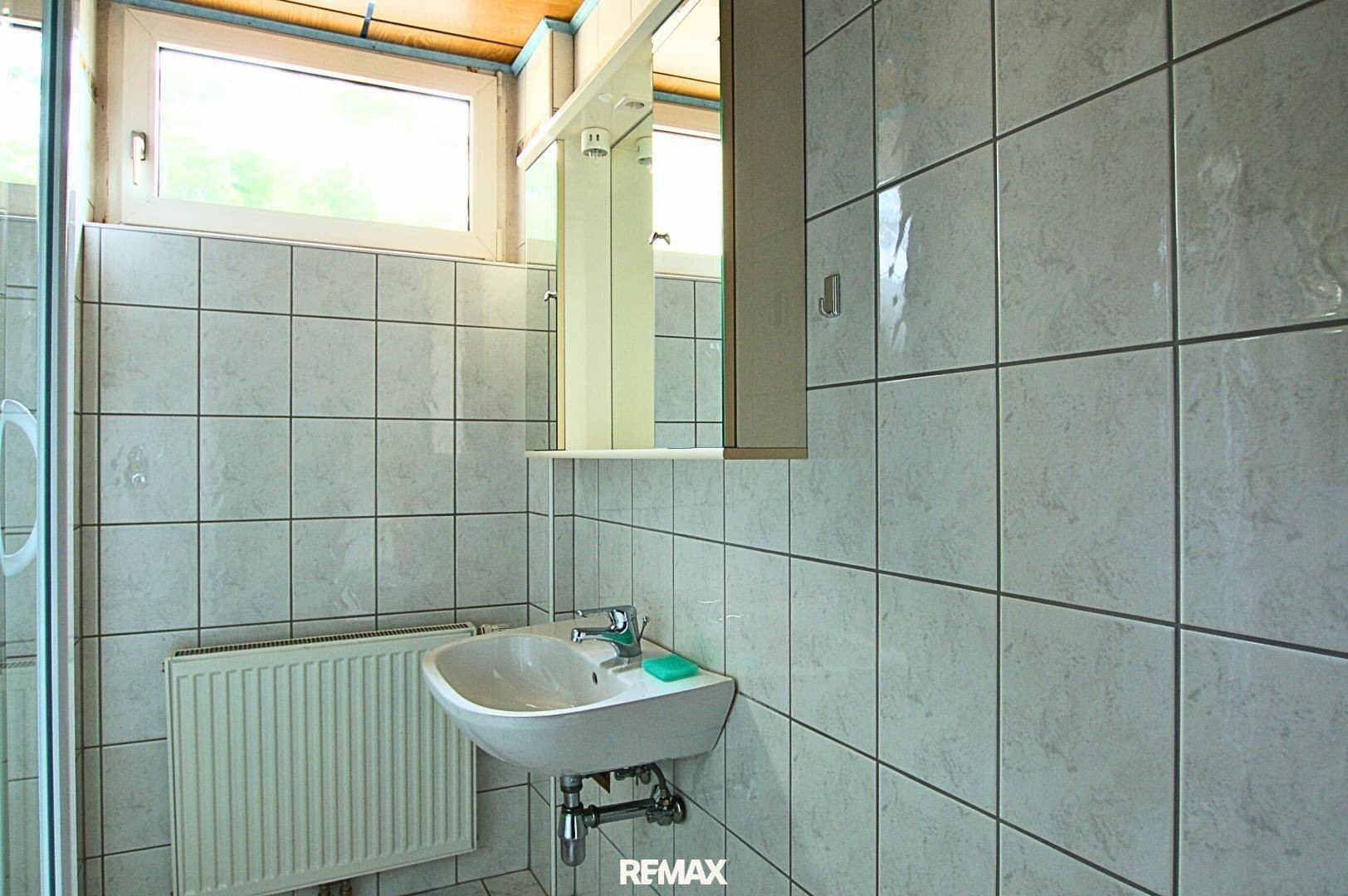 Funktionales Badezimmer mit weißen Fliesen, Waschbecken, Spiegelschrank und Fenster.