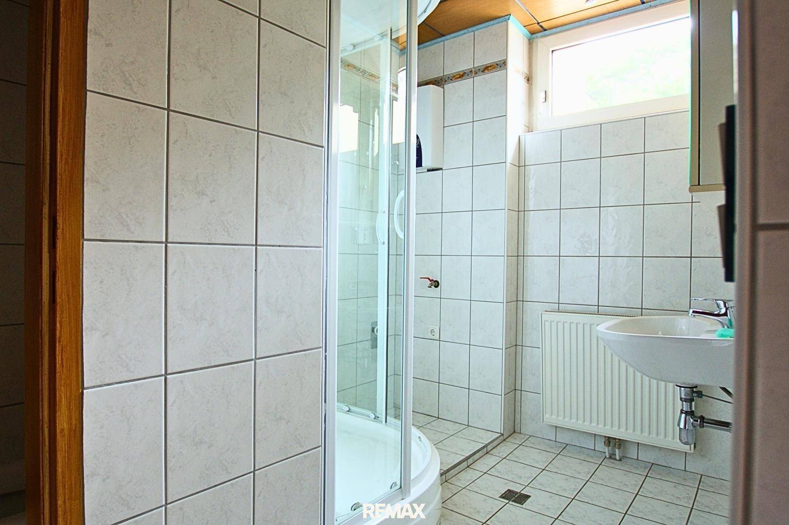 Helles Badezimmer mit weißen Fliesen, Duschkabine, Waschbecken und einem Fenster.