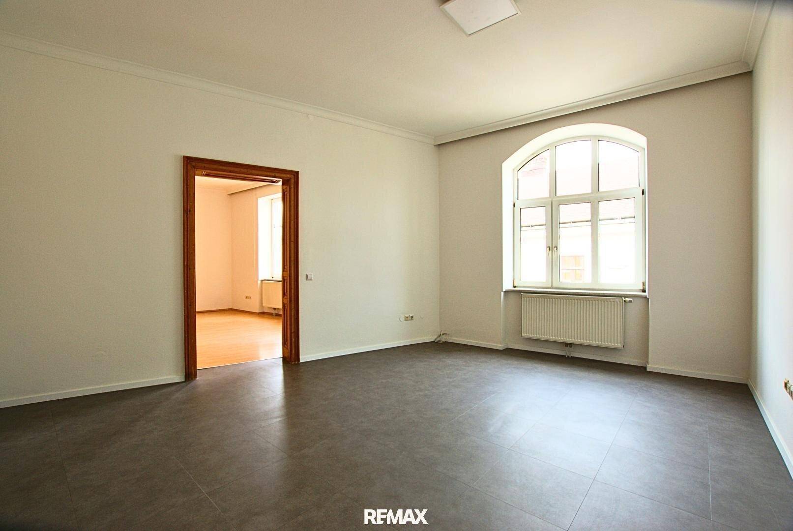 Helles Zimmer mit grauen Fliesenboden, hohen Decken und einem großen Bogenfenster mit Heizkörper.
