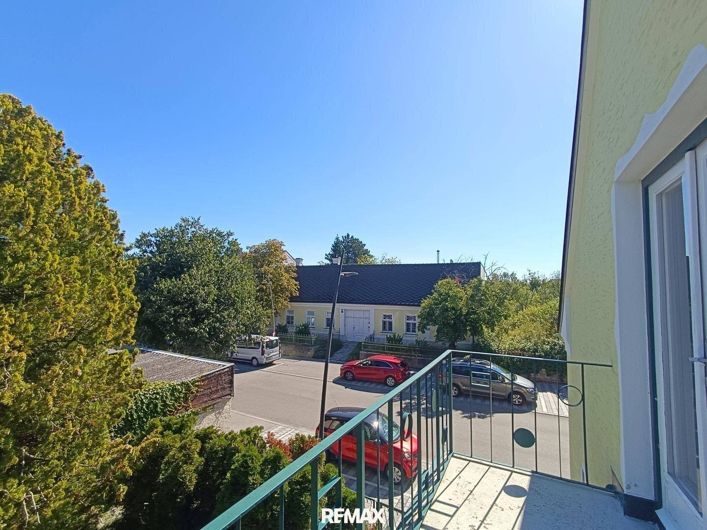 Blick vom Balkon auf die ruhige Straße und umliegende Gebäude mit viel Grün an einem sonnigen Tag.