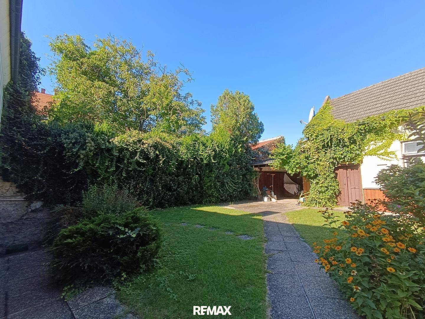 Gepflegter Gartenbereich mit Rasenfläche, dichtem Bewuchs und einem Steinweg unter blauem Himmel.