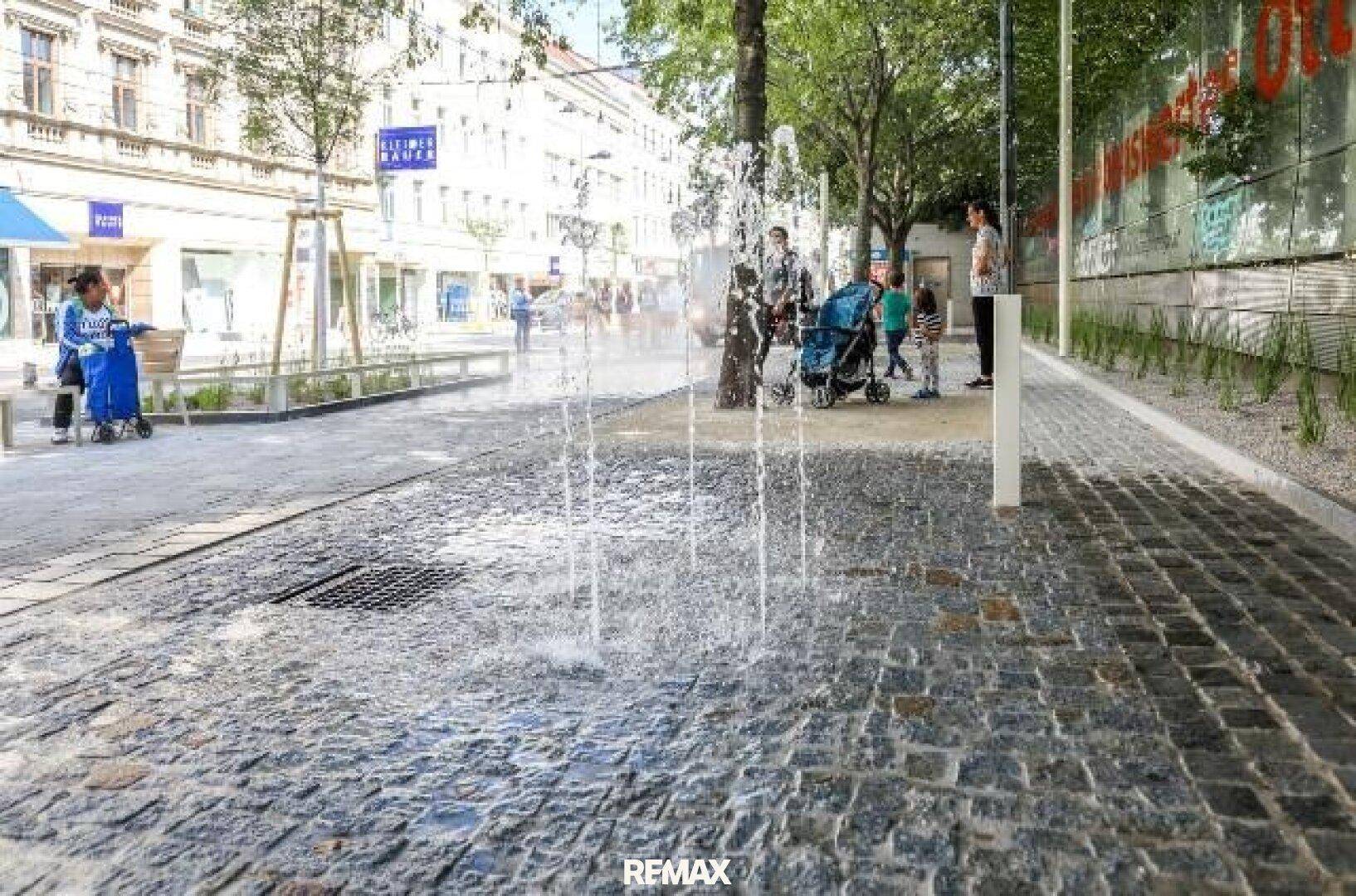 Belebte Stadtstraße mit Kopfsteinpflaster, einem Wasserspiel und Bäumen im Hintergrund.