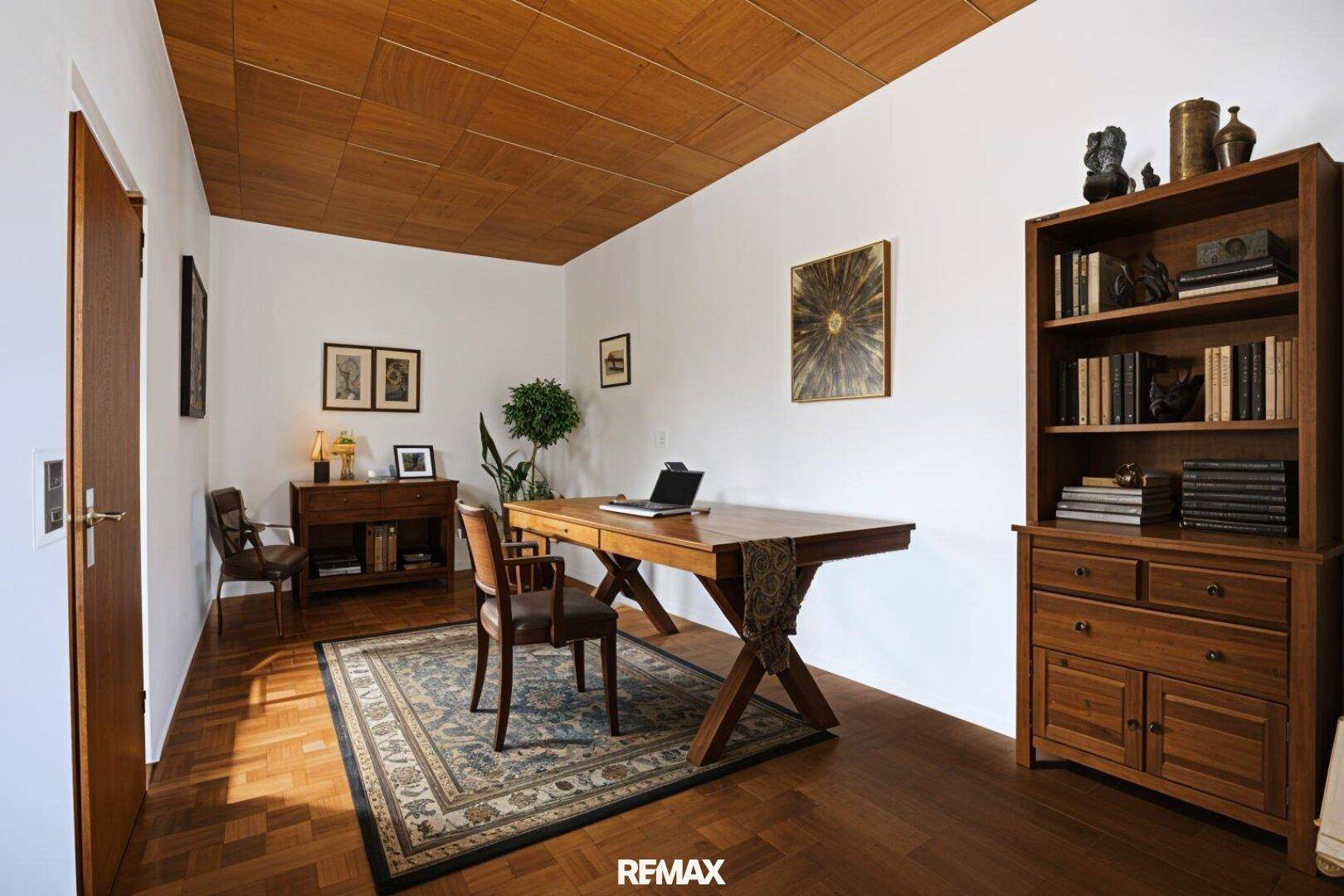 Helles Arbeitszimmer mit großem Holzschreibtisch, Bücherregal und einer Holzdecke.