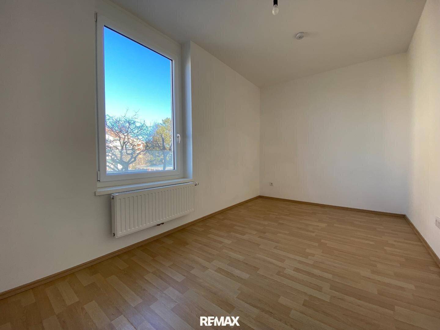 Helles, leeres Zimmer mit Parkettboden und einem großen Fenster, das den Raum erhellt.