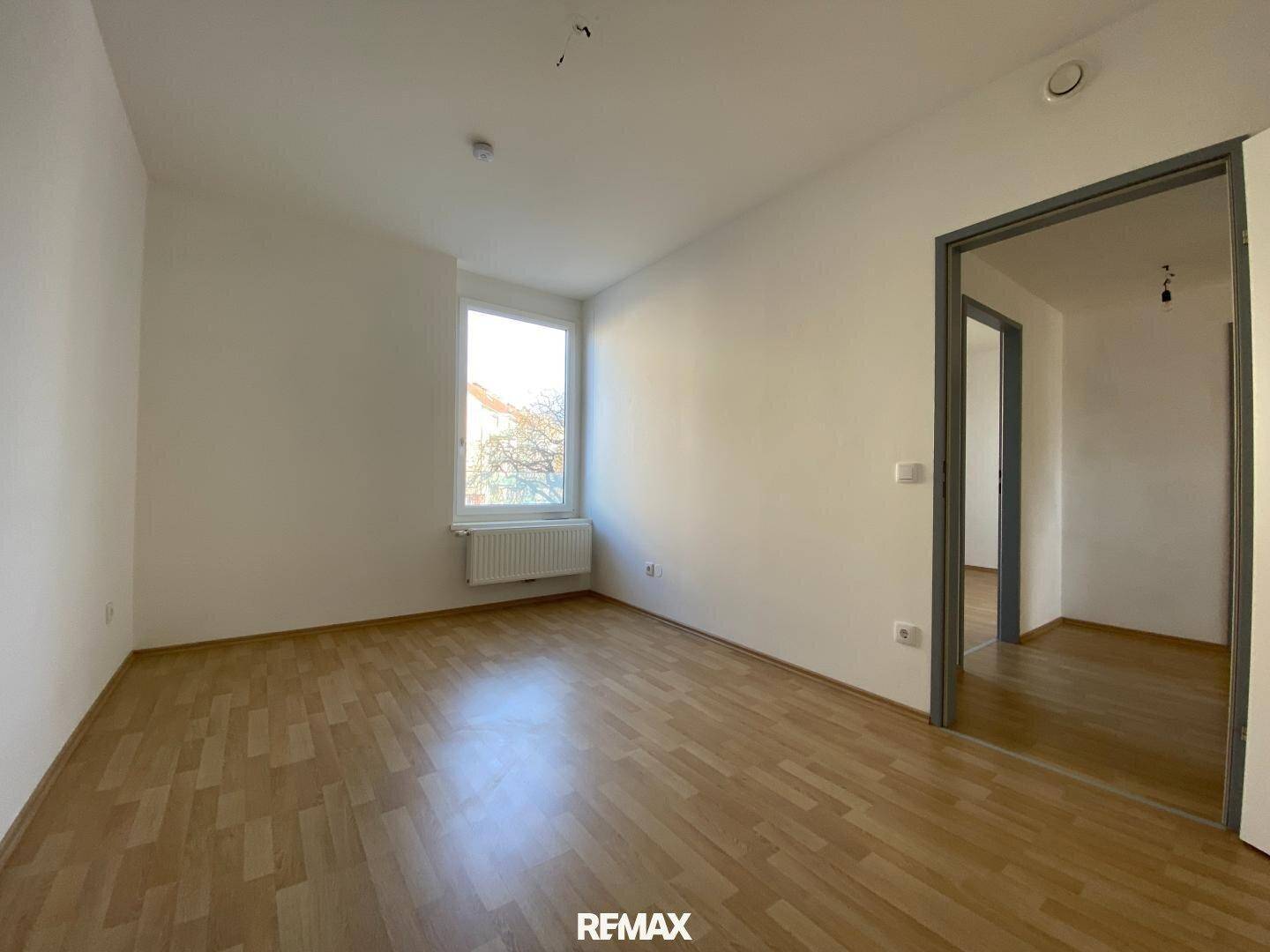 Leeres Zimmer mit Parkettboden und einem Fenster, das viel Tageslicht hereinlässt.