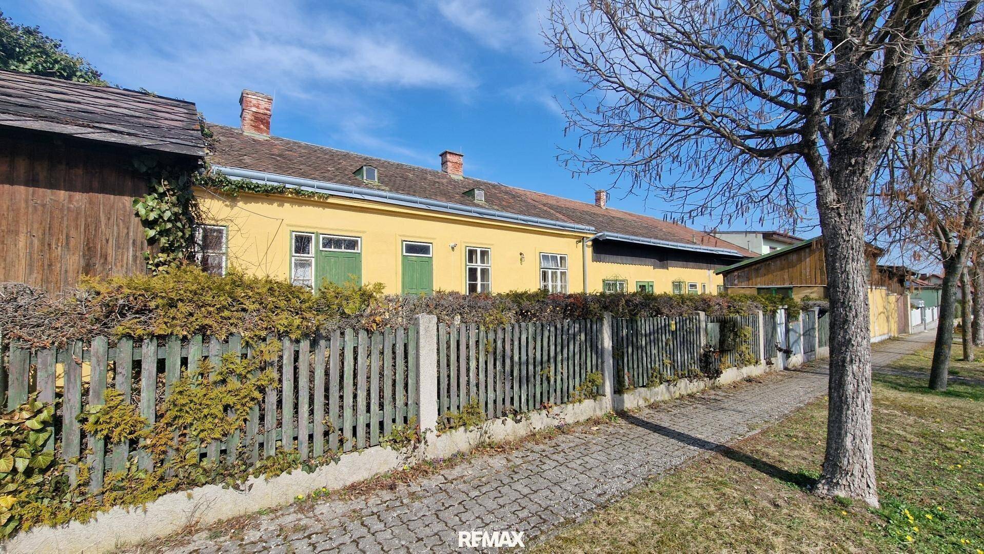 Ansicht des gelben Gebäudes von der Straße mit Holzzaun und Bäumen im Vordergrund.