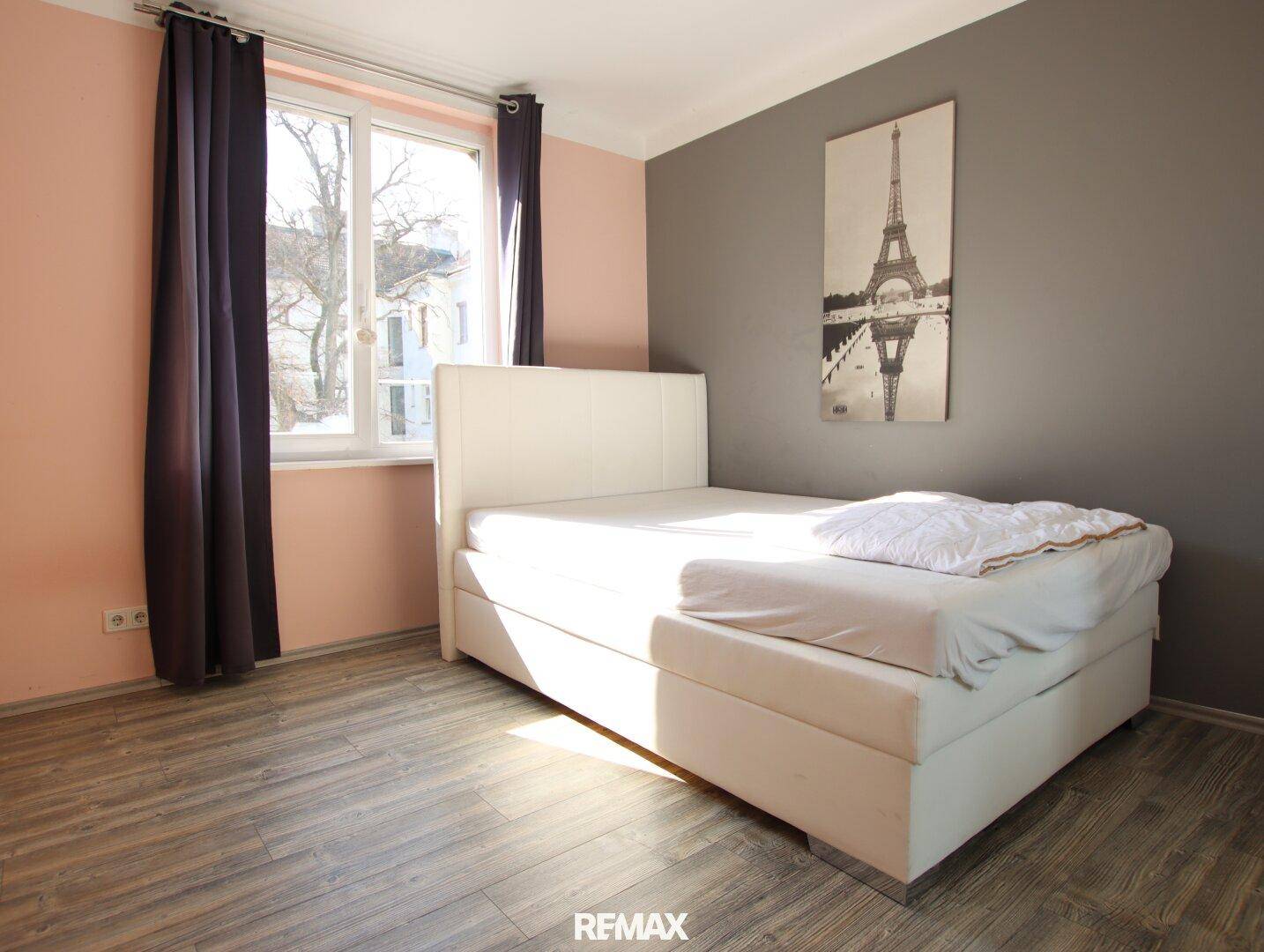 Schlafzimmer mit Doppelbett, Laminatboden und farbigen Wänden, hell durch ein großes Fenster.