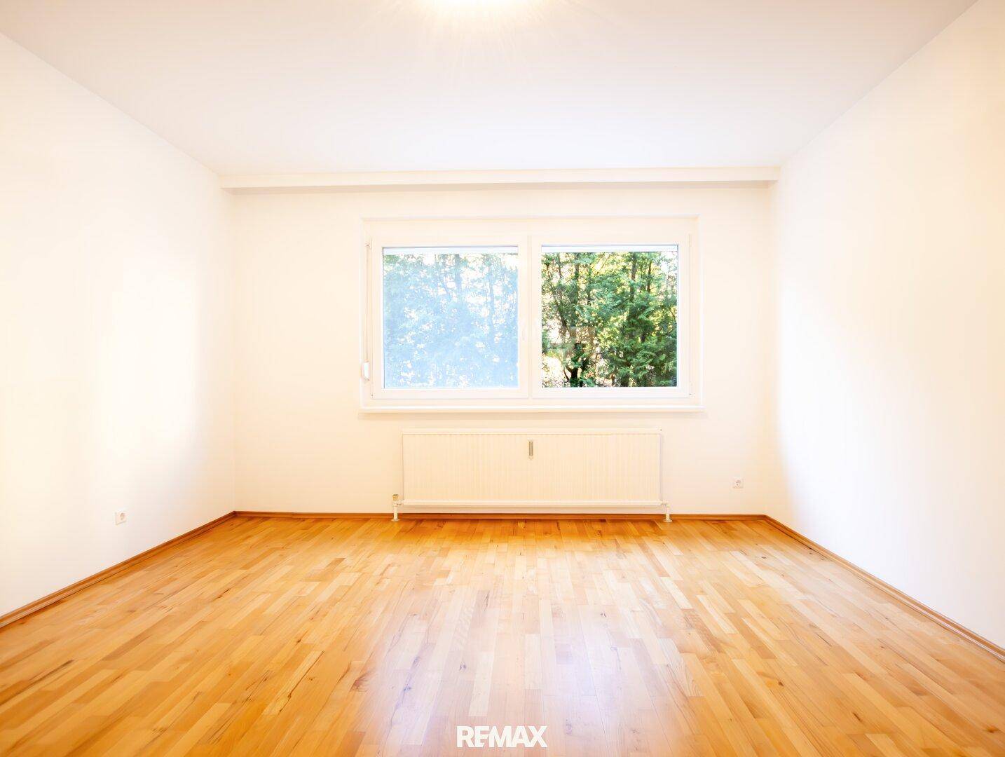 Leeres Schlafzimmer mit hellem Parkettboden und großem Fenster, das viel Tageslicht hereinlässt.