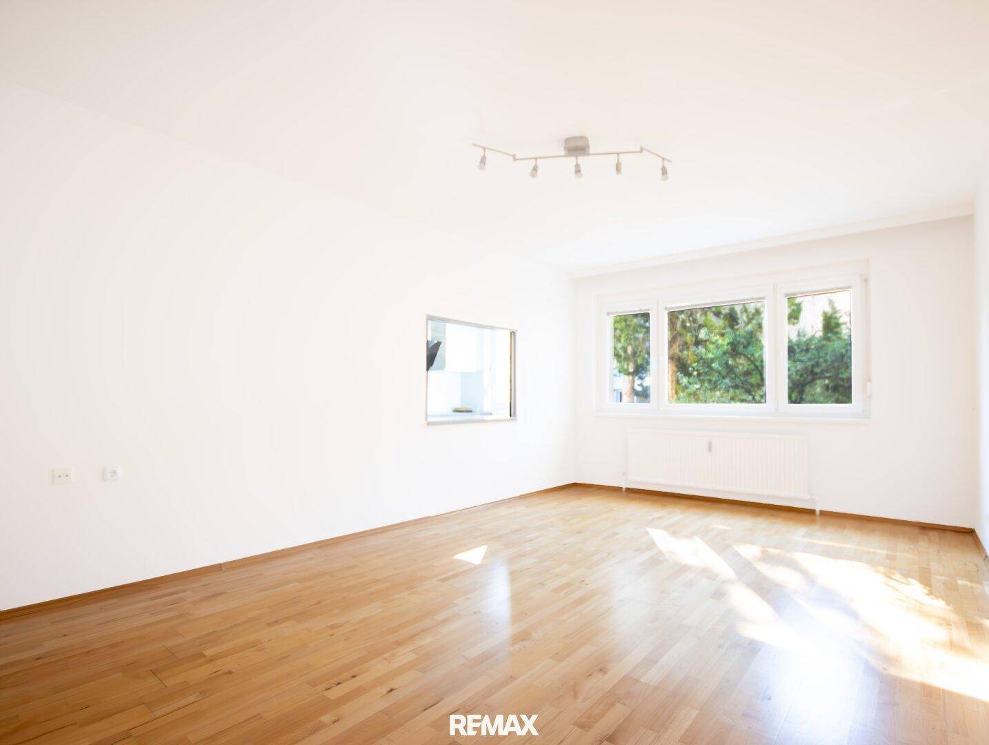 Leeres Wohnzimmer mit hellem Holzfußboden, großen Fenstern und einem Durchgang zur Küche.