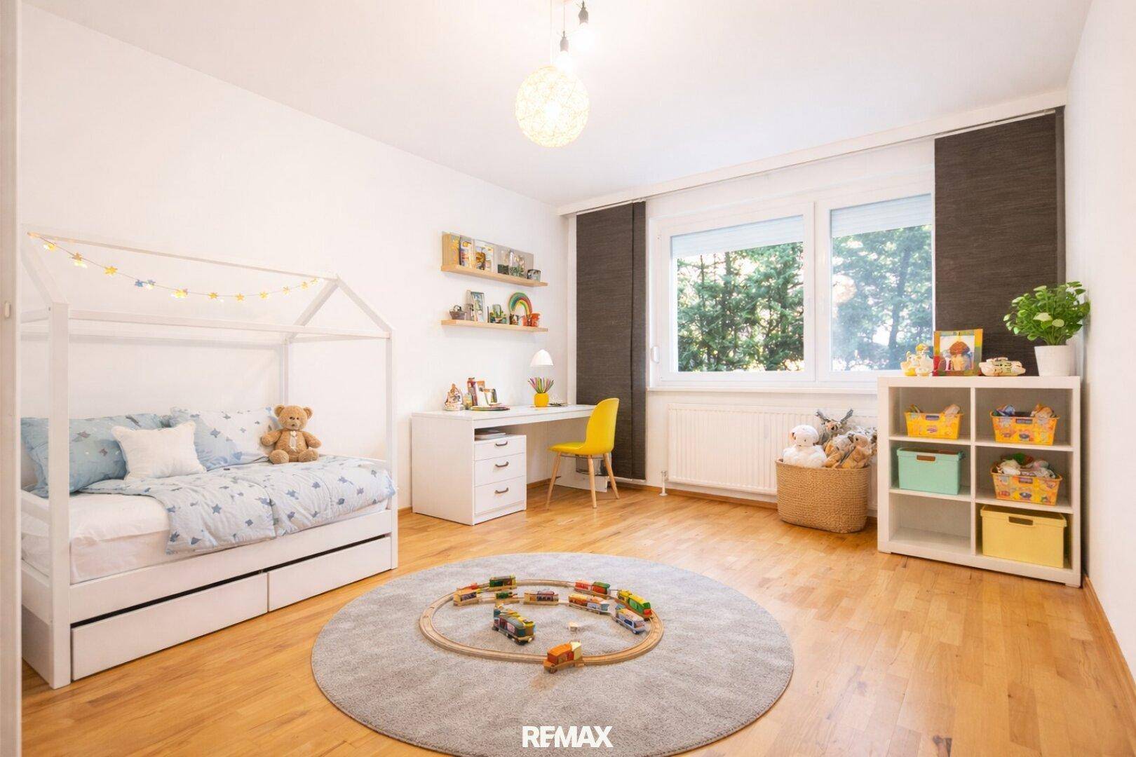 Freundliches Kinderzimmer mit Holzbett, Schreibtisch und Spielzeug, hell durch großes Fenster.