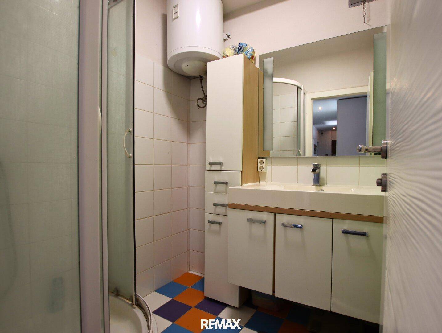 Badezimmer mit Duschkabine, Waschbeckenunterschrank und farbenfrohen Bodenfliesen, praktisch eingerichtet.