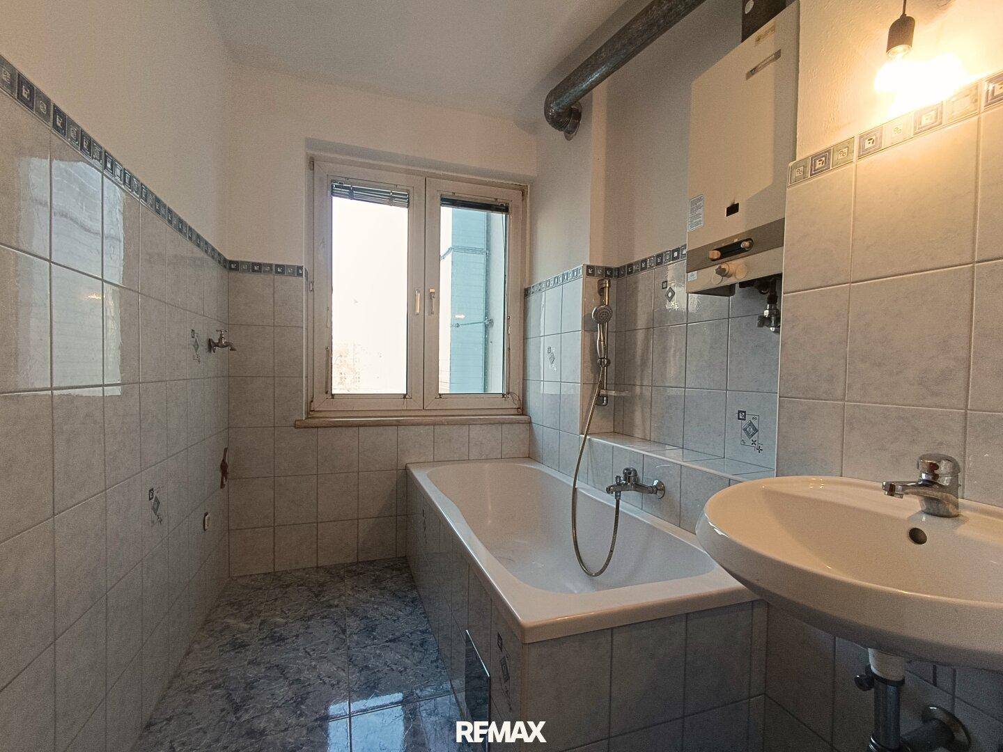 Badezimmer mit Badewanne, Waschbecken, Fenster und hellen Fliesen im 80er-Jahre-Stil.