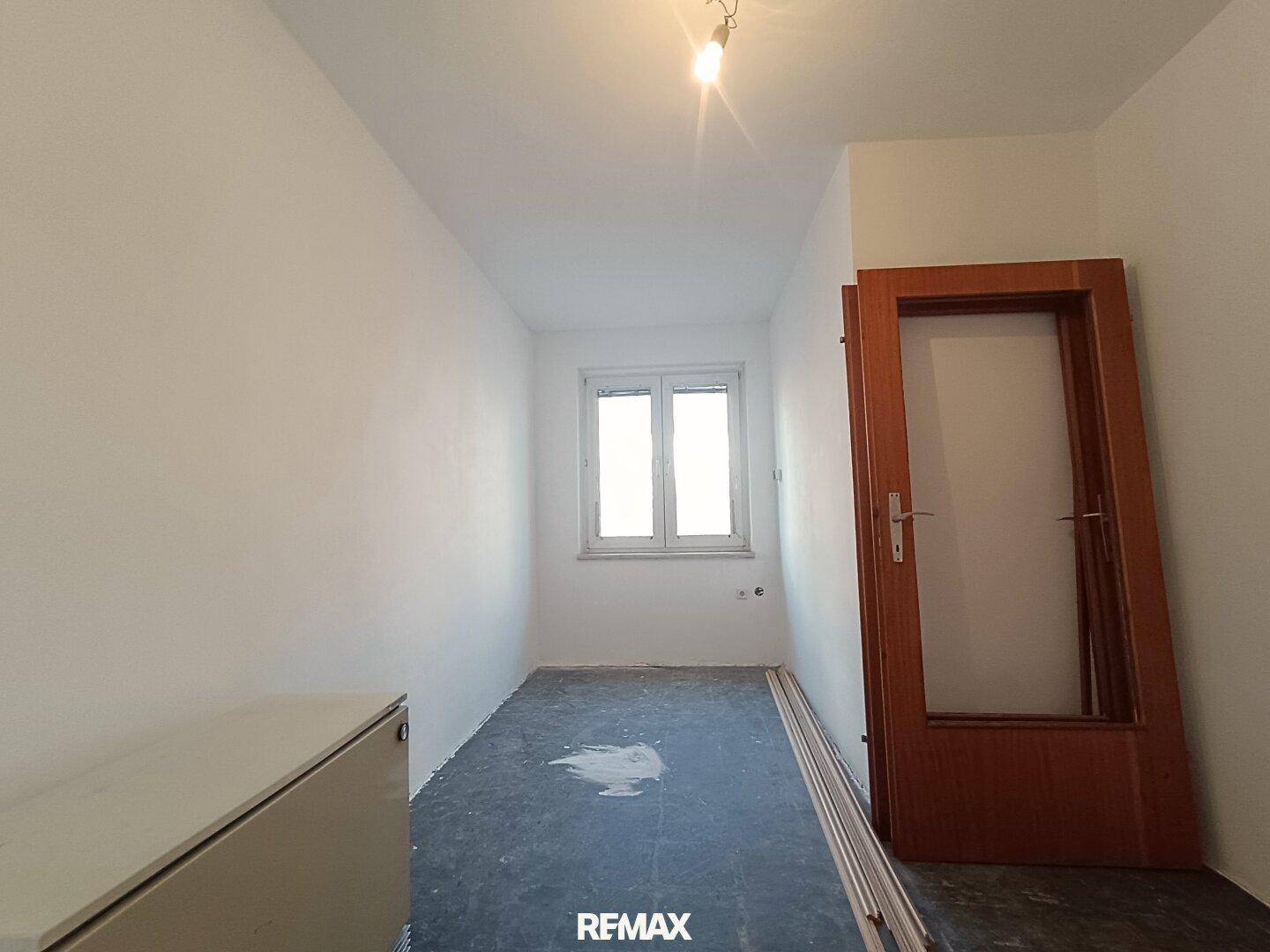 Kleiner Raum mit Fenster, dunklem Boden und einer Holztür, ideal als Abstellraum.
