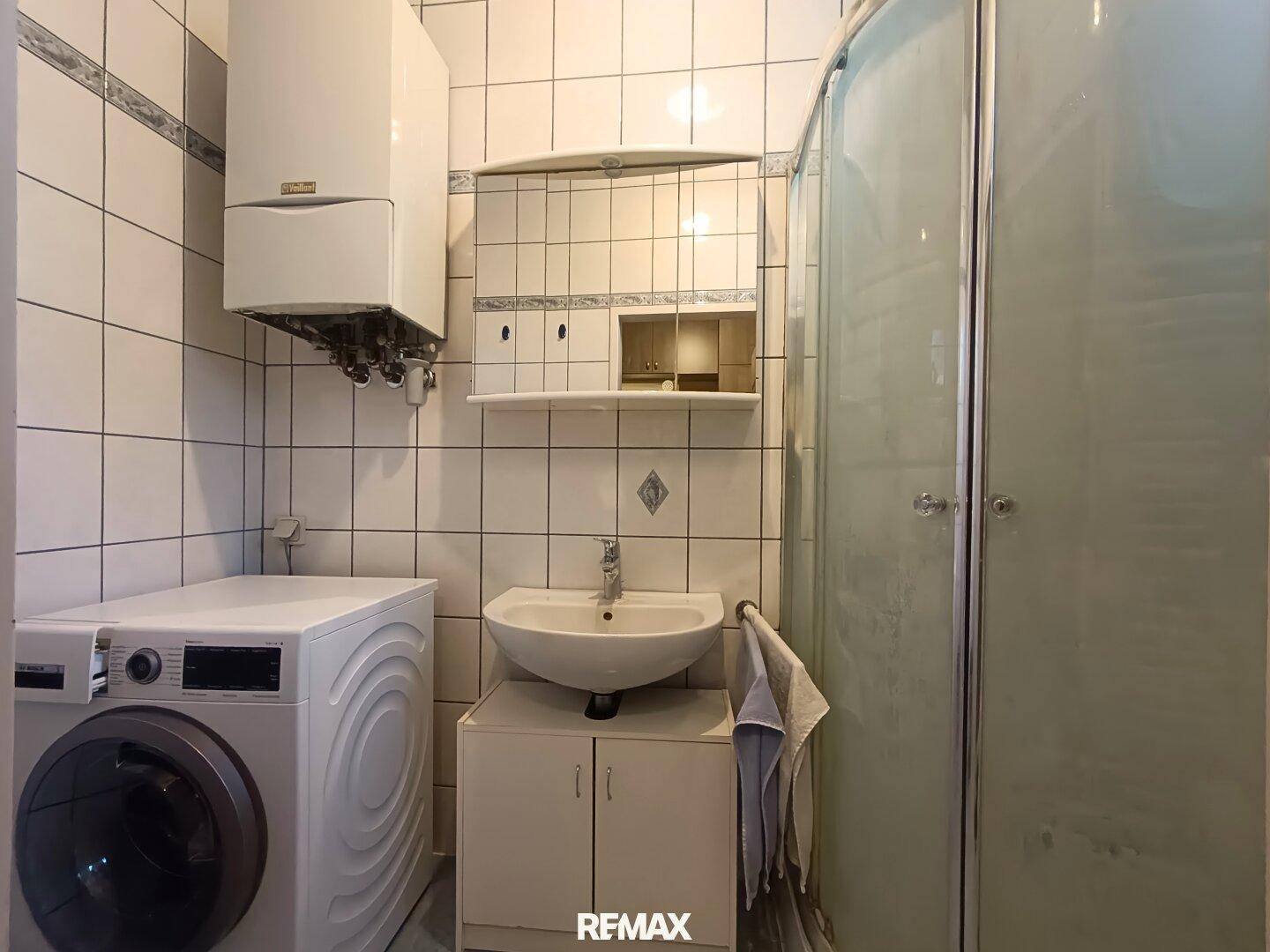 Gefliestes Badezimmer mit Dusche, Waschbecken und Waschmaschine, hell und funktional eingerichtet.