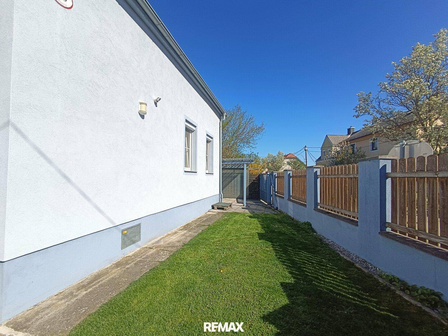 Seitlicher Gartenweg mit Rasenfläche und Blick auf die Hausfassade und den Holzzaun unter blauem Himmel.