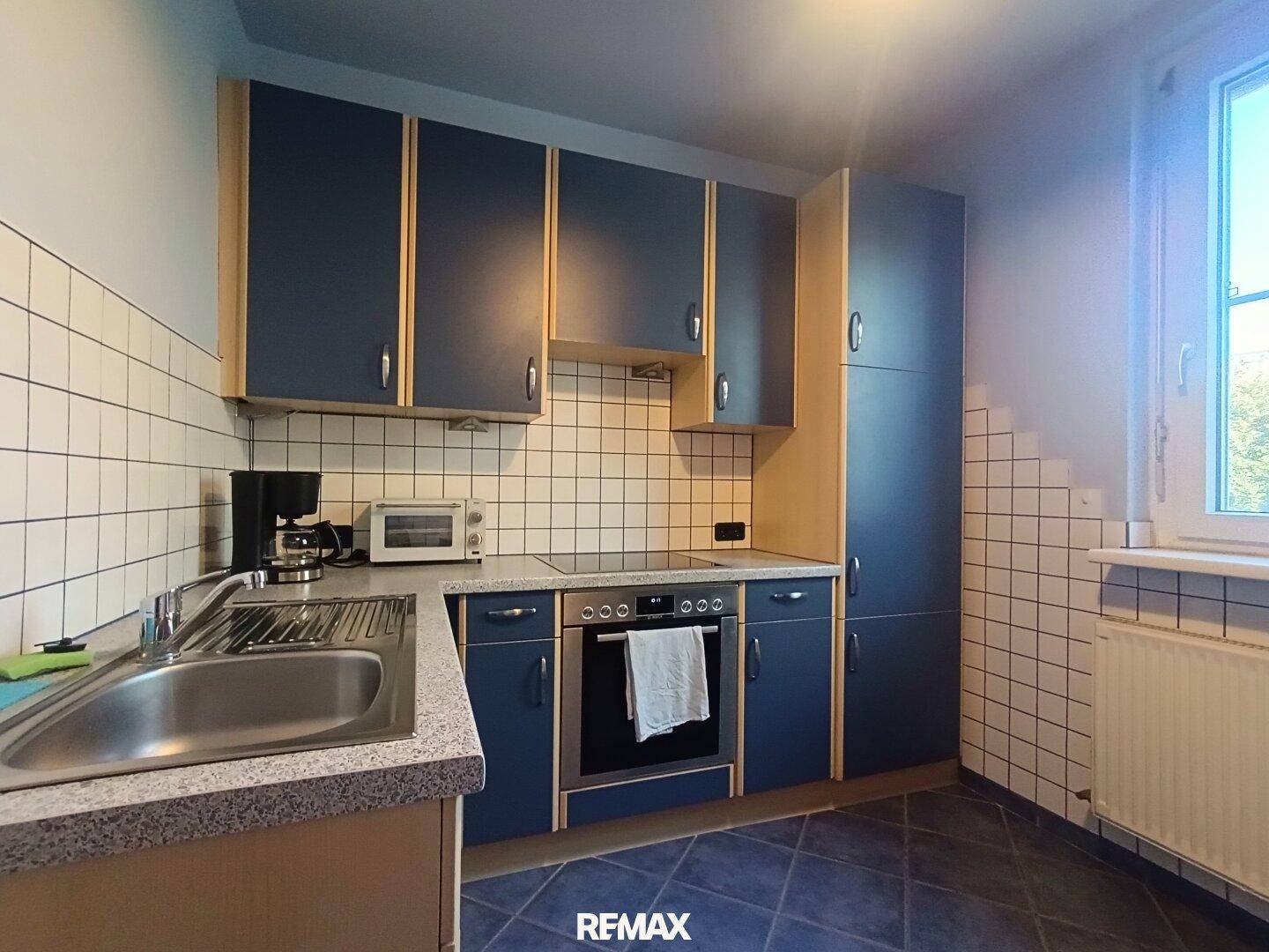 Funktionale Küche mit blauen Schränken, weißen Fliesen und Fenster für Tageslicht, gut ausgestattet.