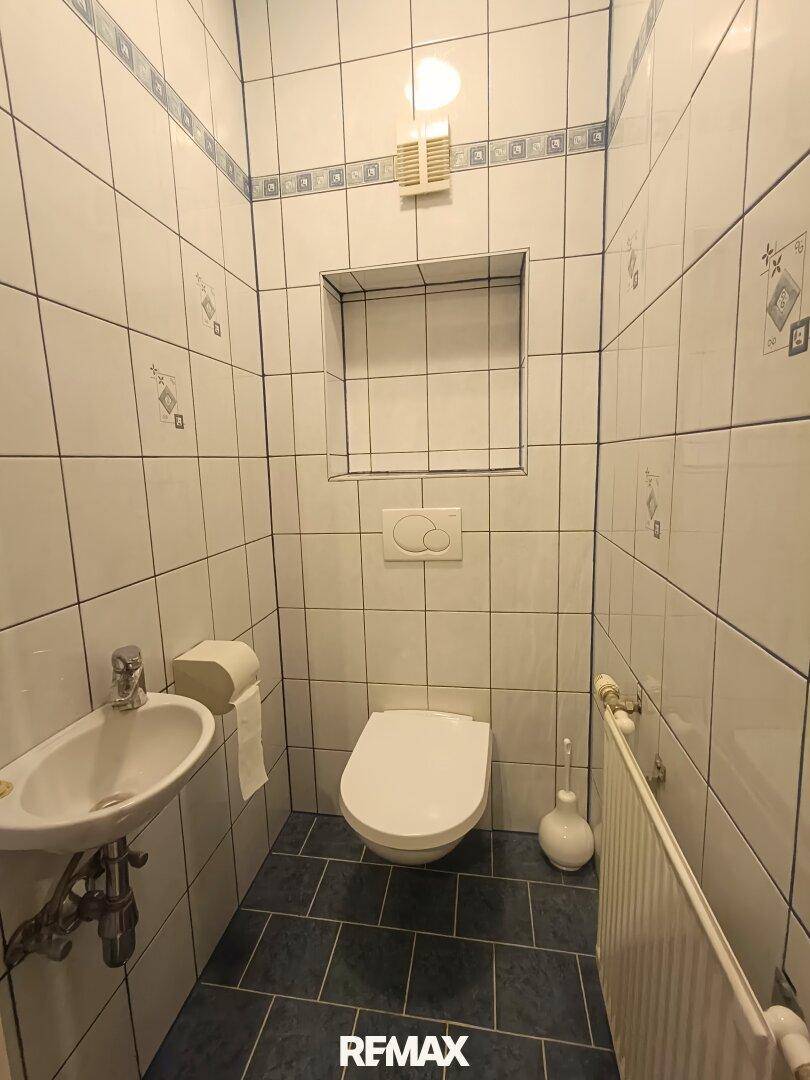 Separates WC mit Wand-WC, kleinem Waschbecken und Fliesen an Wänden und Boden, sauber und funktional.