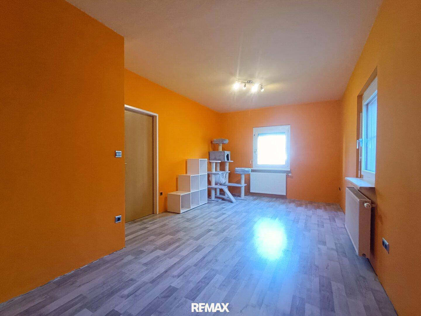 Helles Zimmer mit leuchtend orangefarbenen Wänden, Laminatboden und großem Fenster, vielseitig nutzbar.